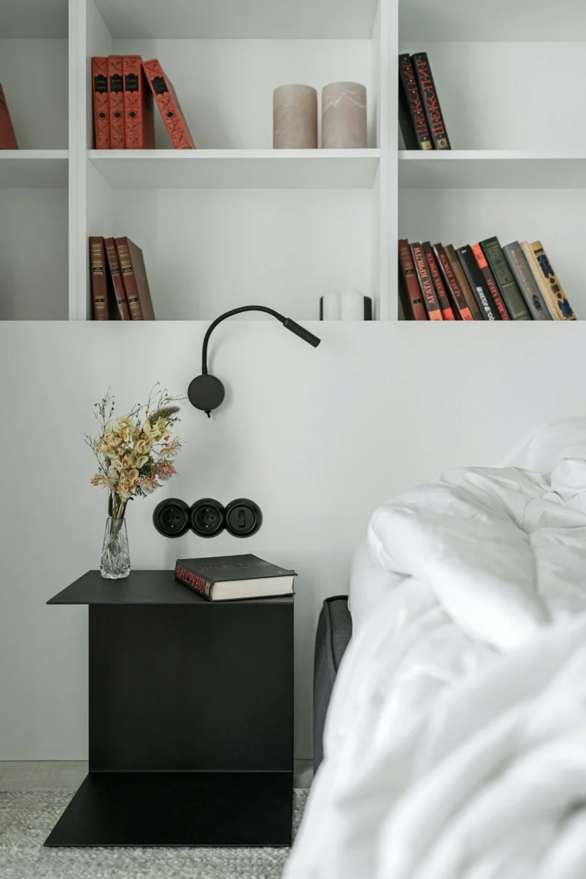 A black bedside table with a book and dried flowers beside a white bed