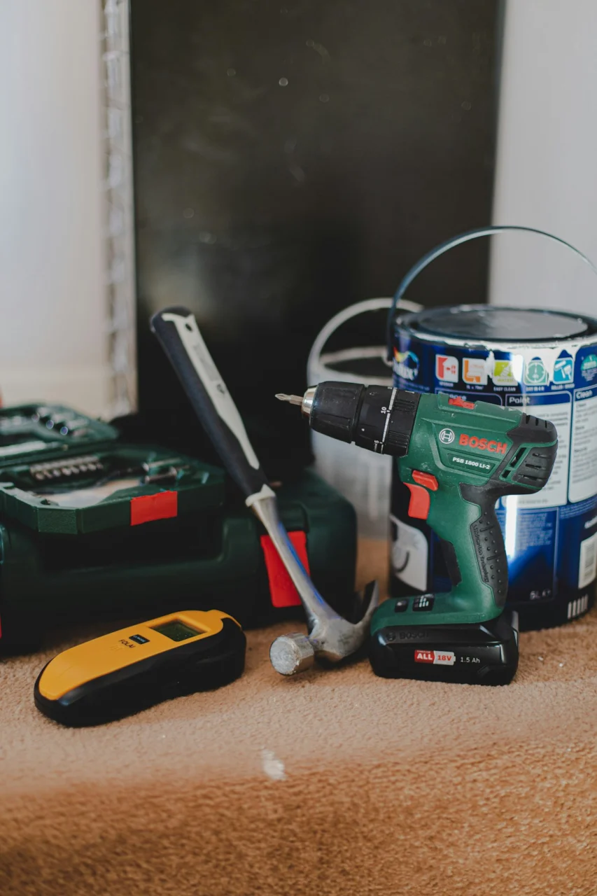 A cordless drill, a hammer, and a yellow stud finder on a carpeted floor