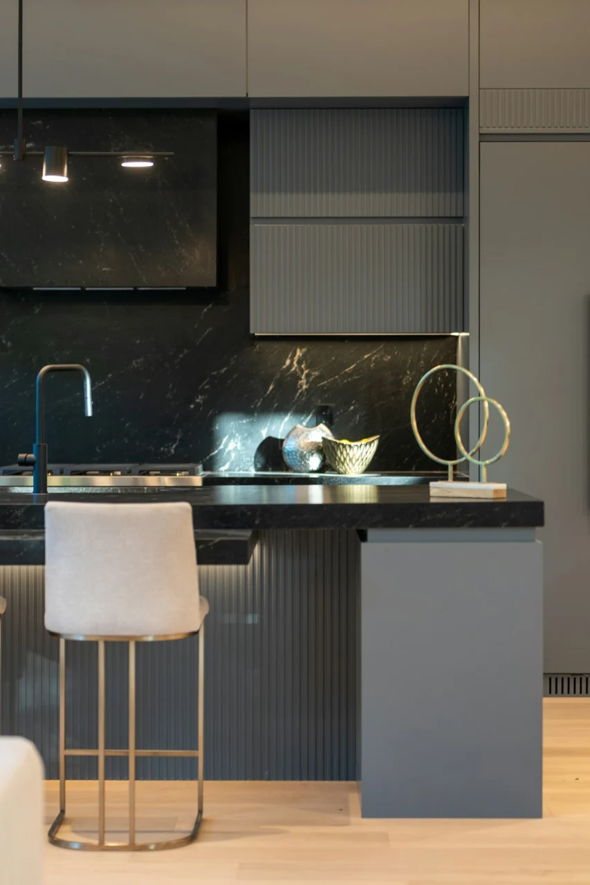 A modern kitchen with dark marble counters, grey cabinets, and a light upholstered stool