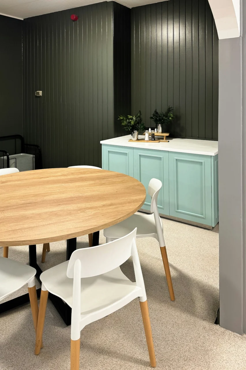 A round wooden dining table with white chairs against a dark green paneled wall
